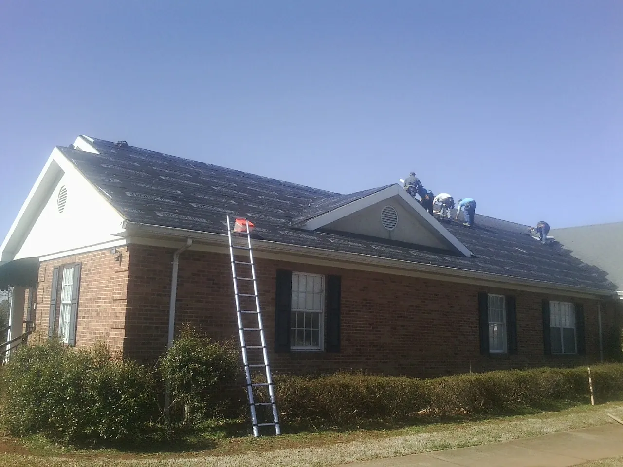 Crew tearing shingles off a brick ranch roof with a ladder set — Emergency Roof Repair work by our crew in Lucile