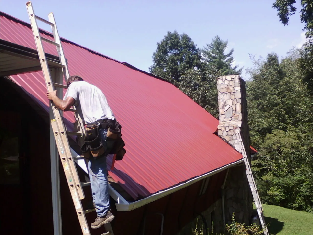 Roofer on a ladder beside a red metal roof and stone chimney — Soffit & Fascia Repair by Roofing Brothers in Lucile